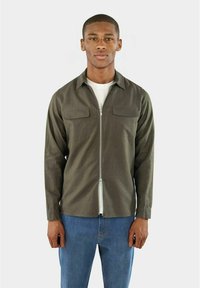 Overhemd met knoopsluiting in donkergroene stof, voorzien van een rits aan de voorkant, twee borstzakken en lange mouwen, gecombineerd met blauwe jeans.