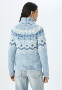 Lichtblauwe gebreide trui met een hoge kraag en geometrische patronen in donkerdere blauwtinten en wit. Ribgebreide manchetten en zoom, zachte textuur.