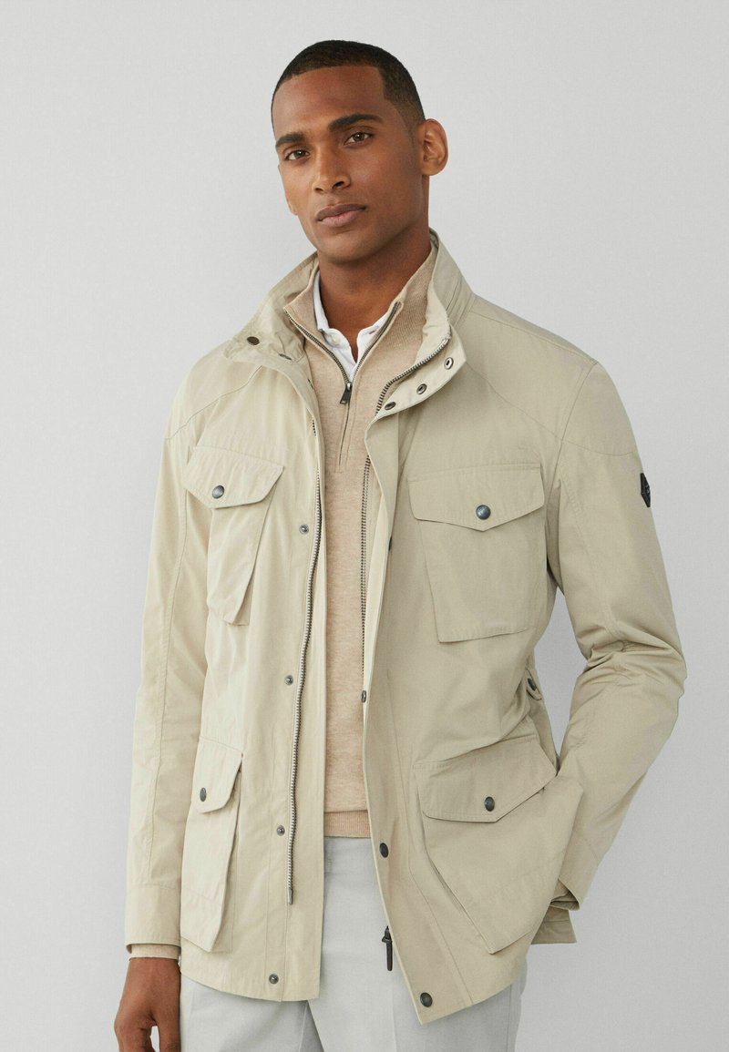 Beige huva jacka med flera framfickor och dragkedjor. Bärs över en lätt tröja och krag skjorta, med en slät, matt textur.