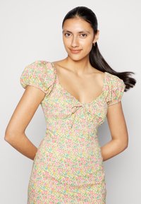 Vestido com estampa floral que apresenta mangas bufantes, decote quadrado e um detalhe de laço na frente. As cores incluem rosa, amarelo e azul.