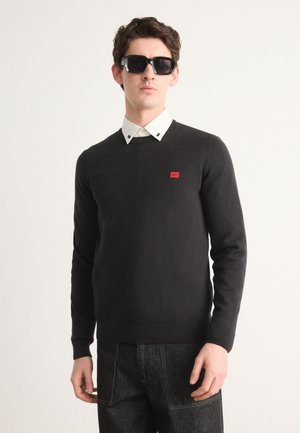 Black sweater with long sleeves, V-neck, and a red branded patch on the chest. Paired with a white collared shirt and dark jeans.
