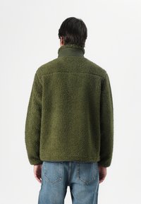 Chaqueta de forro polar verde oliva con superficie texturizada, cuello alto y mangas insertadas, combinada con jeans de mezclilla azul claro.