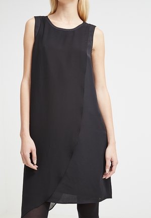 Femme portant une robe noire asymétrique sans manches avec un col rond et un panneau avant superposé, debout devant un fond clair uni.
