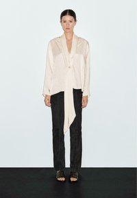 Blusa beige chiaro a maniche lunghe con frontale drappeggiato e dettaglio della cravatta, abbinata a pantaloni neri aderenti e sandali scuri. Tessuto liscio.
