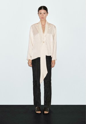 Light beige, long-sleeve blouse with a draped front and tie detail, paired with black, fitted pants and dark sandals. Smooth texture.