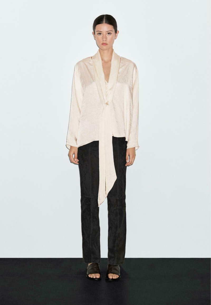 Blusa beige chiaro a maniche lunghe con frontale drappeggiato e dettaglio della cravatta, abbinata a pantaloni neri aderenti e sandali scuri. Tessuto liscio.