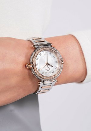 Ezüst és rózsaarany fém órája textúrázott fehér számlappal, díszített órák jelölőivel és fonott kerettel. Kapcsolódik egy láncos karkötőhöz.