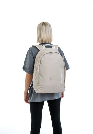 Zaino beige con una texture liscia, forma arrotondata e tasca frontale con zip. Ha spallacci imbottiti e un dettaglio logo discreto.