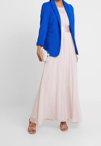 Niebieska teksturowana marynarka nałożona na różową sukienkę maxi z wcięciem w talii. Całość uzupełniają biały clutch ozdobiony perłami i nagie sandały na obcasie.
