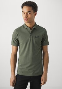 Olivově zelená polo košile vyrobená z jemné bavlny, s klasickým límcem, krátkými rukávy a černým logem na hrudi. Jednoduchý design s kontrastními akcenty.
