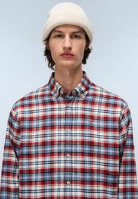 Giovane uomo che indossa un berretto beige e una camicia a scacchi rossa, blu e bianca con bottoni, su uno sfondo grigio chiaro uniforme.