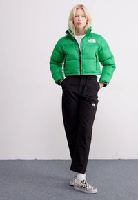 Giacca a vento verde con colletto alto e zip frontale, abbinata a pantaloni neri con cintura. Scarpe slip-on a quadri bianchi e neri.