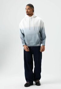 Sweatshirt in wit tot lichtblauw verloop, met een voorvak, trekkoorden en geribde boorden. Gecombineerd met losse, donkergekleurde jeans en zwarte schoenen.