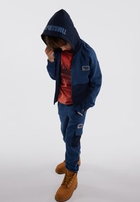Marineblauwe hoodie met logo, rode t-shirt, marineblauwe broek met zakken, en bruine laarzen. Zachte materialen; gelaagd ontwerp met kleurblokken.