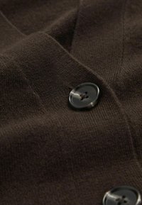 Hnědý pletený cardigan s výrazným černým knoflíkem a texturovanou tkaninou, který působí měkce a útulně.