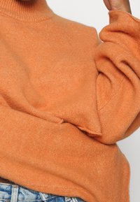 Sweter z golfem, wydziergany z miękkiej, teksturowanej pomarańczowej tkaniny, z opuszczonymi ramionami i luźnymi rękawami, zestawiony z jasnoniebieskimi dżinsami.