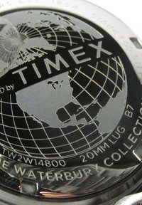 Fond de montre en acier inoxydable avec un design de globe noir, gravé "Timex" et détails du modèle, présentant un travail de lignes complexe et des contours géographiques.