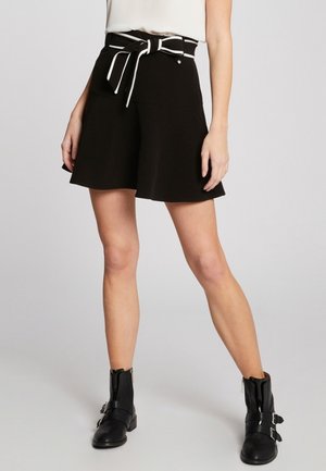 Jupe noire évasée avec une ceinture en tissu blanc à la taille, comportant deux poches avant et une texture lisse. Portée avec des bottines noires.