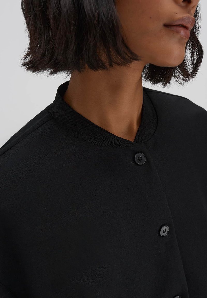 Schwarzes, knöpfbares Kleidungsstück mit rundem Kragen und strukturiertem Stoff. Verfügt über schwarze Knöpfe entlang der Vorderseite und eine glatte Oberfläche.