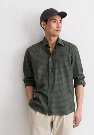 Olive green checkered button-up shirt with a relaxed fit, rolled sleeves, and gray cap; worn with beige corduroy pants.
