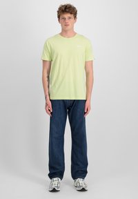 Hellgrünes T-Shirt mit kurzen Ärmeln und Rundhalsausschnitt, kombiniert mit lockeren dunkelblauen Jeans und grauen Turnschuhen. Tattoos an den Armen sichtbar.