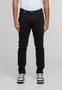 Fekete farmernadrág, slim fit szabással, piros kiemelő címkével, öt zsebbel és gombos záródással, fekete-fehér sportcipőkkel párosítva.