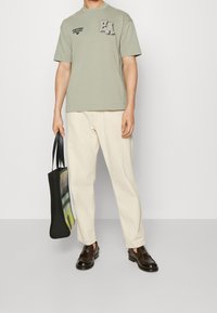 Jasnozielona bawełniana koszulka z czarnymi nadrukami logo, kremowe spodnie, ciemnobrązowe loafersy i czarna torba na ramię z abstrakcyjnym wzorem.