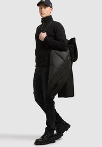 Černá fleecová mikina na zip, černá péřová bunda přes rameno, štíhlé černé džíny a robustní černé boty. Jednoduchý design, bez vzorů nebo ozdob.