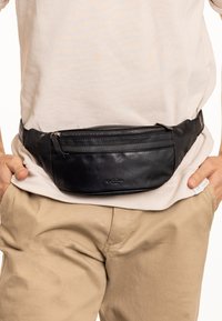 Marsupio in pelle nera con tasca anteriore con zip e texture liscia, indossato in vita sopra una camicia chiara e pantaloni beige.