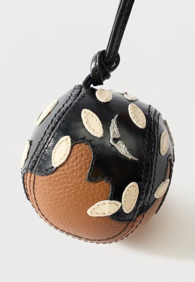 Στρογγυλό μαύρο και καφέ δερμάτινο μπρελόκ με λευκά ραμμένα οβάλ πατς και ένα μικρό μεταλλικό έμβλημα φτερού, προσαρτημένο σε μαύρο δερμάτινο λουρί.
