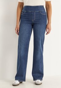 Vrouw in blauwe hoge taille wijde jeans en witte sneakers, staand tegen een effen lichte achtergrond.