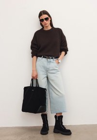 Naine kannab tumedat pruuni bleiserit, heledat sinist laiade säärega teksapüksi, musti pitsitud saapaid ja prille, hoides musta kott-poolikuga valge seina ees.