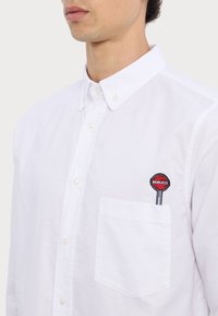 White button-up shirt with a collar, featuring a red and black Fiorucci logo embroidered on the chest pocket. Textured fabric, standard fit.