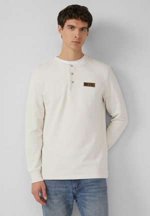 Cremfarbendes Langarmshirt mit Rundhalsausschnitt, drei-Knopf-Leiste und rechteckigem Brustpatch. Getragen mit blauen Jeans.