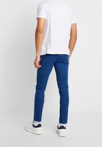 Modré elastické kalhoty, slim fit design, viditelné švy, v kombinaci s bílým tričkem s krátkým rukávem a bílými teniskami s námořnickými akcenty.