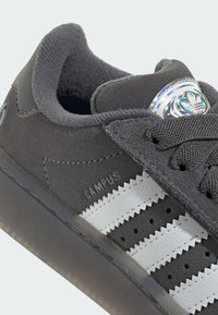 Harmaat mokkanahkakengät, joissa on kolme valkoista nahkaviivaa, tekstuurilla varustetut lenkkarit ja pyöreä heijastava yksityiskohta kielekkeessä.