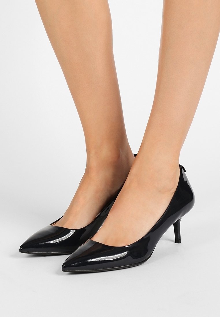Jambes portant des escarpins noirs vernis à bouts pointus à talons kitten sur un fond neutre.