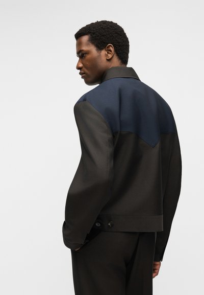 Άνδρας με σκοτεινό χρώμα δέρματος, που φοράει μαύρο μπουφάν με μπλε σκούρο μέρος στους ώμους, σε στάση τριών τετάρτων πλάτης σε απλό φόντο.