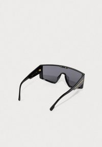 Store svarte oversized skjoldsolbriller med mørke linser og slanke stenger, vist fra en bakre vinkel på en lys bakgrunn.