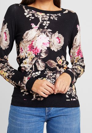 Vrouw draagt een zwart gebreide trui met lange mouwen met grote beige, roze en gele bloemmotieven, gecombineerd met een blauwe spijkerbroek.