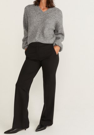 Suéter gris de punto con cuello en V, combinado con pantalones negros de corte a medida. El conjunto presenta un ajuste relajado y texturas suaves. Incluye tacones negros puntiagudos.