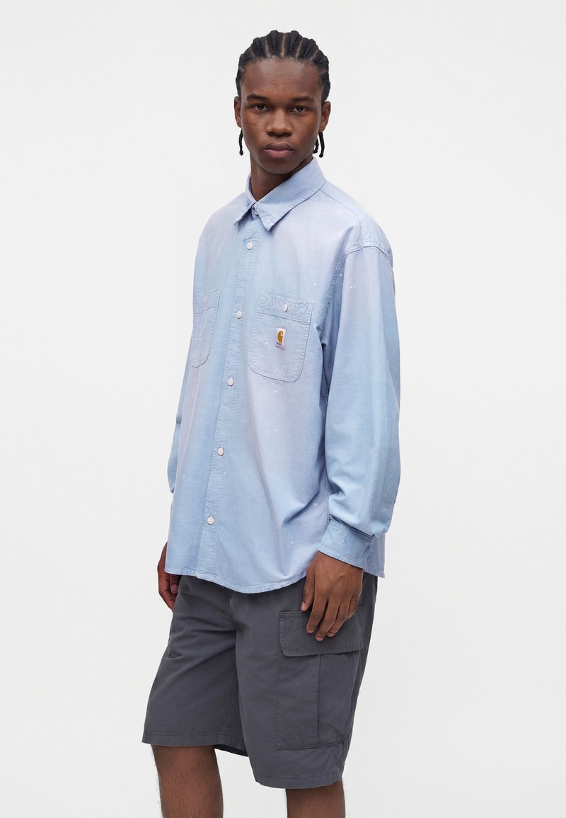 Giovane uomo con capelli intrecciati, indossa una camicia azzurro chiaro con bottoni e pantaloncini cargo grigio scuro su uno sfondo bianco uniforme.