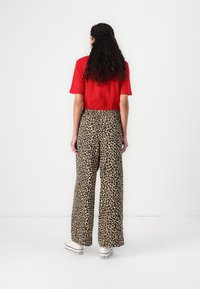 Top rosso a maniche corte abbinato a pantaloni a gamba larga con stampa leopardata beige e nera; il modello indossa sneaker bianche. Sfondo minimalista.