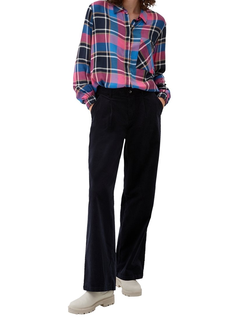 Femme portant une chemise à carreaux multicolore, un pantalon noir taille haute à jambes larges, des bottines blanches à semelles épaisses, les mains dans les poches.
