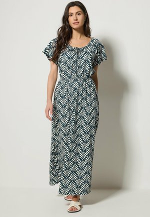 Frau in einem dunkelgrünen Maxikleid mit weißem Blattmuster, kurzen Ärmeln, elastischer Taille und weißen Sandalen, die nach vorne geht.