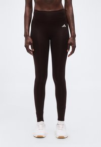 Άτομο που φοράει μαύρο κολάν Adidas με ψηλή μέση και λευκά αθλητικά παπούτσια, στέκεται μπροστά σε απλό φωτεινό φόντο.