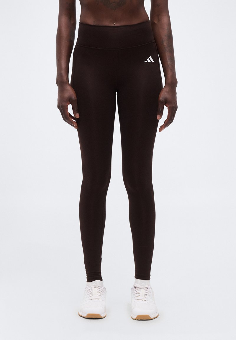 Άτομο που φοράει μαύρο κολάν Adidas με ψηλή μέση και λευκά αθλητικά παπούτσια, στέκεται μπροστά σε απλό φωτεινό φόντο.