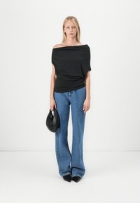 Zwarte off-the-shoulder top met gedrapeerd ontwerp, gecombineerd met lichtblauwe wijde jeans en een kleine zwarte handtas. Textuurdetails van de stof zijn zichtbaar.