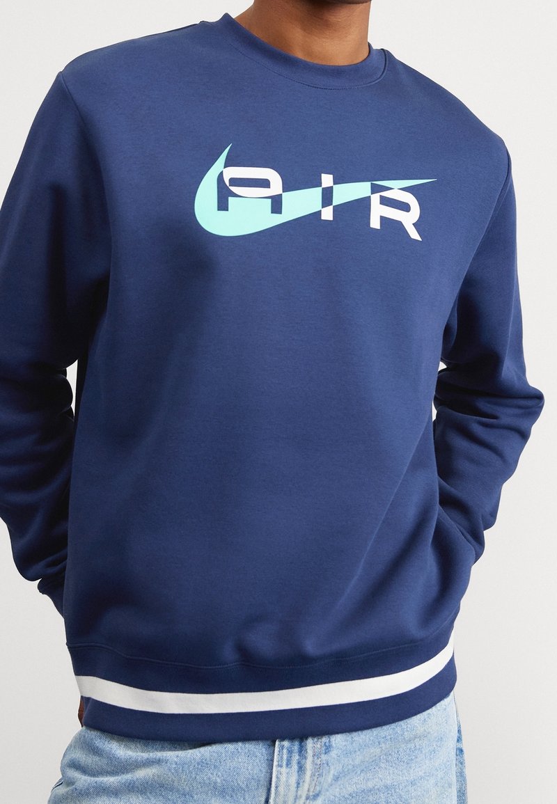 Mörkblå sweatshirt med en ljusblå Nike-logotyp och vit text "AIR." Ribbstickade muddar och fåll med vita ränder ger detaljer.