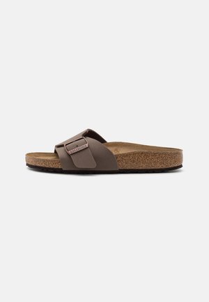 Brown slide sandal with a wide strap and adjustable buckle, cork footbed, and textured rubber sole. Minimalist design, lightweight material.